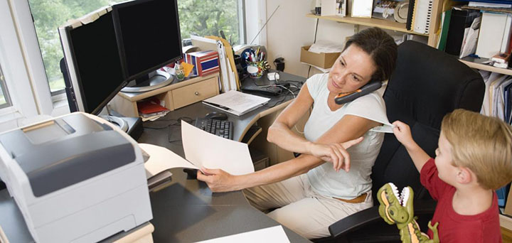 A mother on the phone handling a print job and gesturing to her son to hold on a minute while seated at a desk.