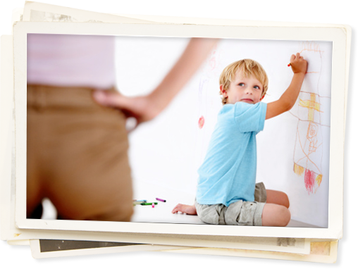 Boy drawing with crayons on wall with parent looking on with hands on hips.